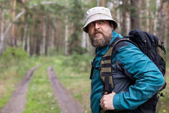 A man with a beard and a hat is standing in a forest