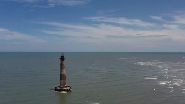 Aerial view of Morris Island Lighthouse standing tall amidst the ocean's embrace, waves creating patterns on the water, Folly Beach, South Carolina, United States.