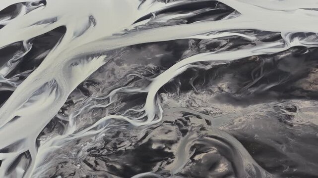 Aerial view of a glacial river system creates abstract patterns of stark white water against the dark, rugged landscape, Iceland.