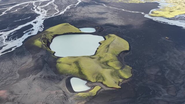 Aerial view of green hills surrounding milky blue lakes amid stark black volcanic sand flats and winding glacial rivers, highlands, Iceland.