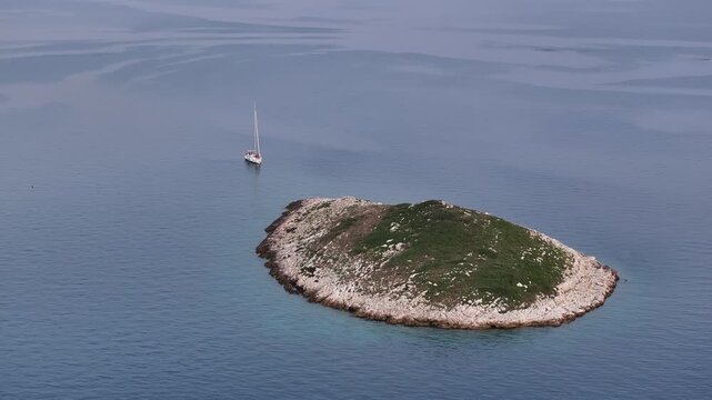 Aerial view of a rocky islet with green vegetation and a single sailboat in tranquil, clear blue waters, Biograd na Moru, Zadar County, Croatia.