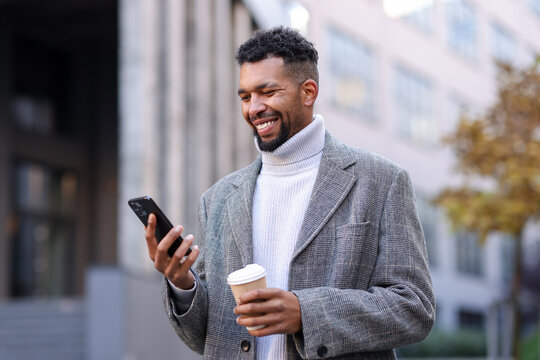 Smiling man in suit with smartphone and takeaway cup on city street, low angle view