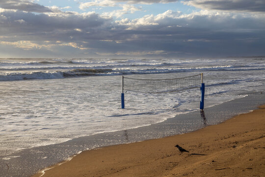 Powerful storm waves cover a beach under sunny skies, with volleyball poles standing in the surf and a crow on the sand. A dramatic scene of nature's force at a Mediterranean resort in low season.

