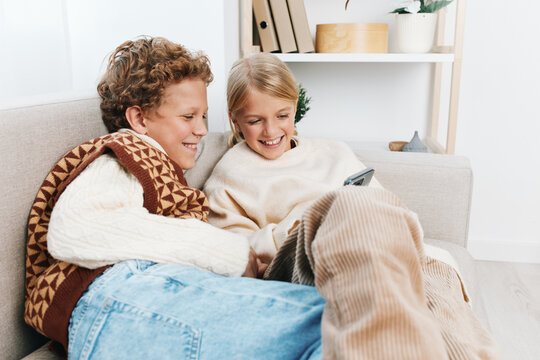 Two happy children sit close on a cozy sofa, wrapped in a warm blanket, sharing a tablet and smiles. Soft natural light highlights a relaxed home moment.