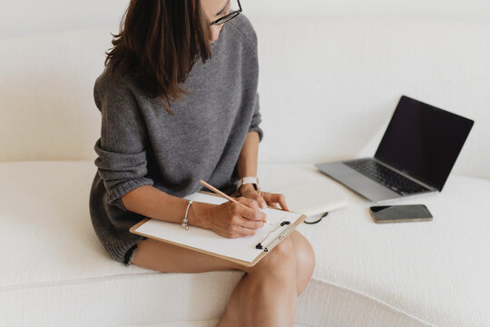 A woman sitting on a white sofa writing on a clipboard, with a laptop and notebook next to her.