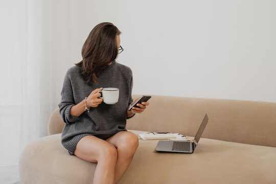 A woman sitting on a sofa in a minimalist bright room, holding a coffee mug and looking at her smartphone while her laptop rests nearby. - Powered by Adobe