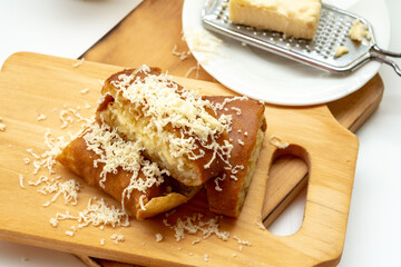 Overhead shot of Indonesian Martabak Manis (sweet pancake) topped with generous grated cheese and butter on a wooden cutting board, with a grater and cheese block visible in the background.