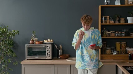Modern gen Z teenager wearing tie-dyed short-sleeved hoodie standing at kitchen, eating sandwich and scrolling news feed on smartphone