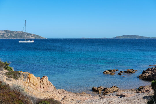 Rocky coastline with clear turquoise water, scattered stones, sparse coastal vegetation, and a sailboat anchored near distant islands under a clear blue sky.