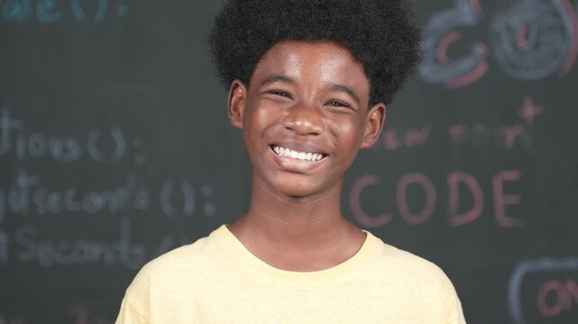 African adult smiling to camera while standing at blackboard with engineering code or prompt. Closeup of handsome teenager wearing casual cloth while attend in STEM technology class. Edification