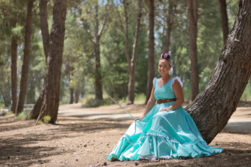 Fototapeta premium Young, beautiful, blonde woman wearing a typical Andalusian flamenco dress in light blue for going to a pilgrimage, holding a stick in her hand, sitting and resting on the trunk of a tree.