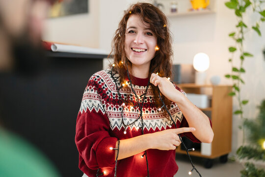 A young woman in a festive sweater smiles as she decorates with string lights, creating a warm and inviting atmosphere for the holidays.