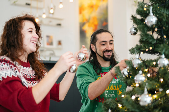 A happy couple decorates a Christmas tree together, adding silver ornaments and string lights to the festive branches.
