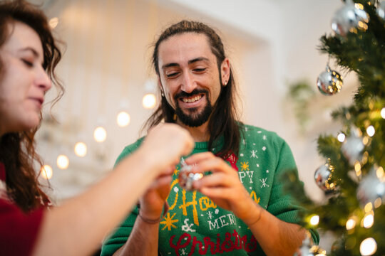 A smiling man in a festive sweater and a woman decorate a Christmas tree with ornaments, surrounded by warm, blurred lights.