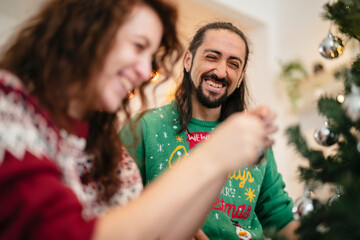 A joyful couple in festive sweaters decorates a Christmas tree together, sharing laughter and holiday cheer.