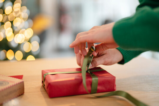 A person's hands tie a green ribbon around a red gift box on a wooden table, with a blurred Christmas tree in the background. - Powered by Adobe