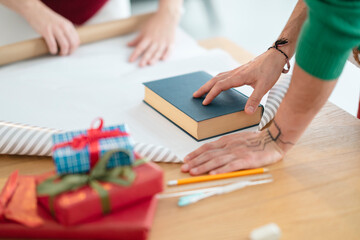 Two people are preparing gifts on a wooden table, with wrapping paper, ribbon, and a book ready for use.