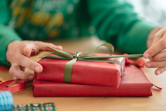 A person in a green sweater is carefully tying a ribbon on a red gift box, preparing presents for a special occasion.