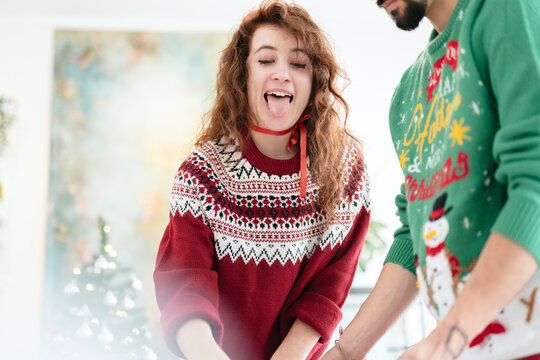 A young woman with curly hair sticks her tongue out playfully while wearing a festive sweater. A man in a similar sweater stands beside her.