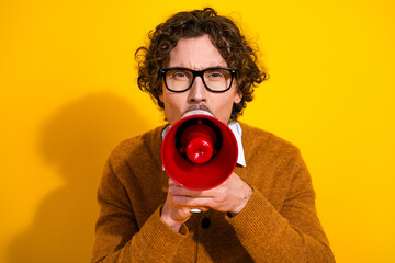 Obraz premium Young stylish man with glasses in a cardigan speaks through a red megaphone against a bright yellow background