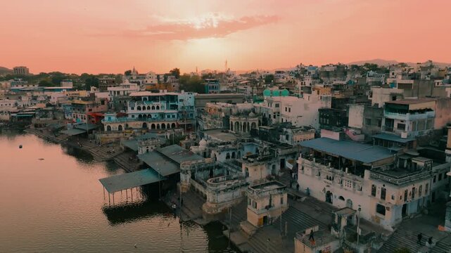 Aerial view of buildings with domes, flat roofs, and trees, set against a backdrop of mountains under a peach-colored sky, Pushkar, Rajasthan, india.