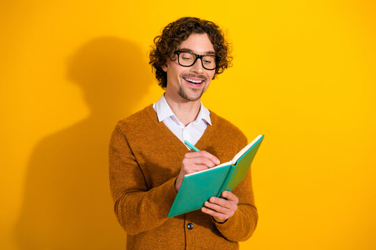 Young man with curly hair in a brown cardigan writes in a turquoise notebook against a bright yellow background - Powered by Adobe