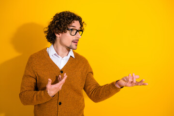 Young stylish man in mustard cardigan speaks with open hands against a yellow background
