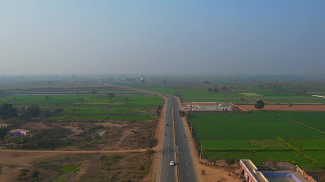 Aerial view of a long, gray road cutting through vibrant green fields and earthy brown patches under a hazy sky, Nankana Sahib, Punjab, Pakistan.