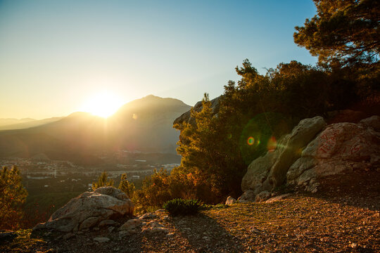 Warm sunset light spilling over rocky hills and forested mountains, creating a soft, glowing, atmospheric landscape filled with evening calm.