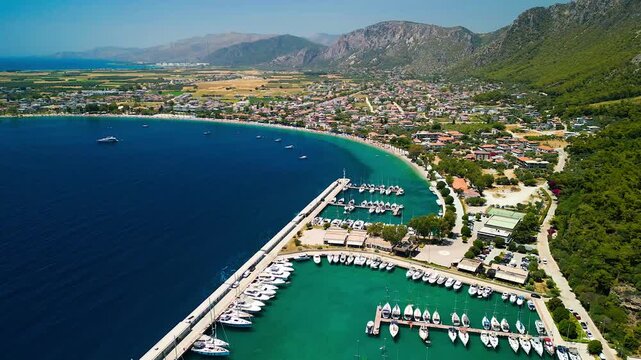 Aerial view of boats docked at Oren Marina, the turquoise sea contrasts with the golden beach and green hills, Oren, Mugla, Turkiye.