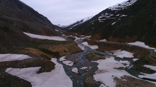 Aerial view of the river snaking through the valley, flanked by snow-dusted mountains, creating a stunning contrast of white and dark rock, Naran, Khyber Pakhtunkhwa, Pakistan.