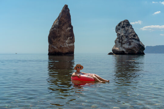 Woman, ocean, beach. Relaxed woman floats on pink ring in clear sea near rocks. Summer vacation, copy space.