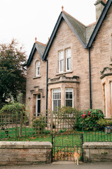 Obraz premium Traditional stone home with gabled roof, bay window, and front garden behind wrought iron gate.