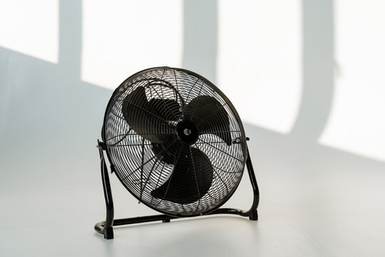 black metal fan on floor, retro industrial tabletop design casting angular shadows across white studio surface; ideal for workshop cooling prop, artisan and photographer set dressing