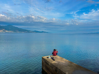Tranquil scene of a fisherman angling on a pier with breathtaking ocean and mountain views, perfect for travel ads