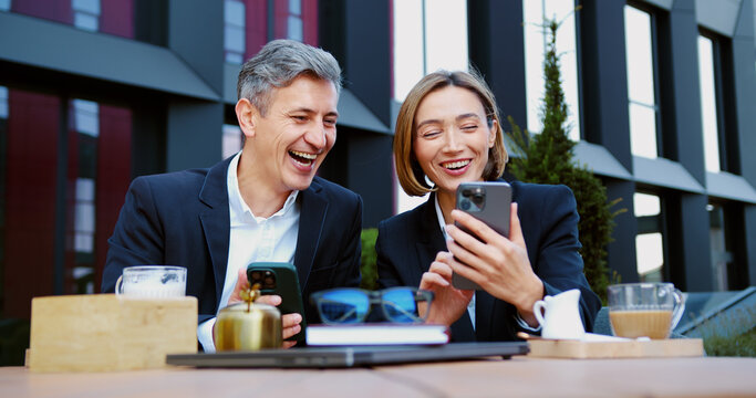 Man and woman in suits having fun, laughing, watching something funny on smartphone during lunch break of the cafe. Business partners smiling and interacting while using mobile phone.