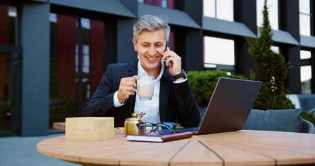 Businessman using smartphone during business call while sipping coffee and typing on laptop. Outdoor workspace, leisure, modern, relaxed, communication, productivity, natural light