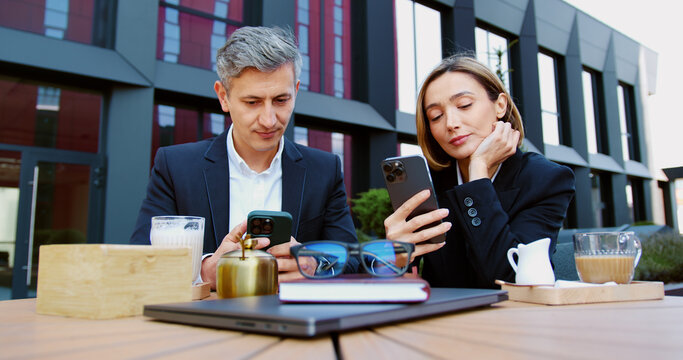 Sad disinterested business man and woman looking disappointedly at uninteresting information on their smartphones and drinking coffee during lunch break sitting on the summer terrace of the cafe.