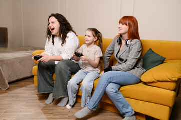 Happy family spends free time playing video games. Mom and daughter play with video game controller while sitting on living room sofa, enjoying eSports, competing, having fun while playing video games