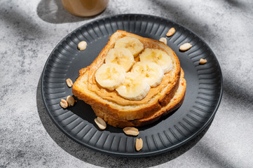 Peanut butter toast and banana on a black plate on a gray background with nuts