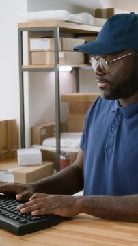 Medium vertical shot of African American male logistics company associate, dressed in blue uniform tshirt and cap, working on computer while filling out consignment forms for parcel shipment