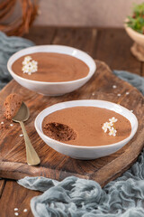 Individual servings of rich chocolate mousse in white bowls, one with a spoonful lifted, garnished with delicate white flowers, presented on a rustic wooden board with soft blue fabric
