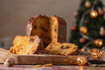 Close-up of golden Panettone slices dusted with powdered sugar and raisins on a wooden board, with a festive Christmas tree and warm bokeh lights in the background