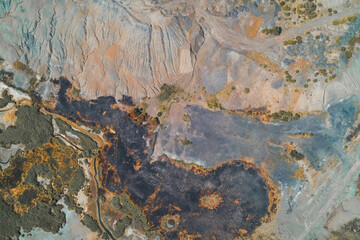 Top-down aerial photograph showing a landscape damaged by fire and industrial runoff. Sharp contrast between black, scorched earth and eroded spoil heap texture.