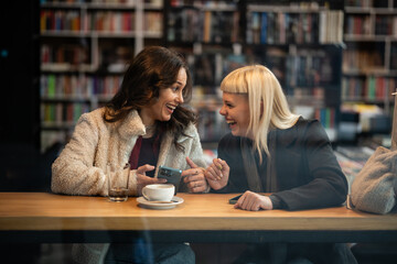 Two women friends laughing in library cafe using smartphone