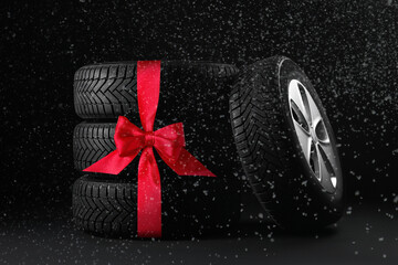 Car wheel and tires with red ribbon bow under falling snow on black background. Special offer or discount