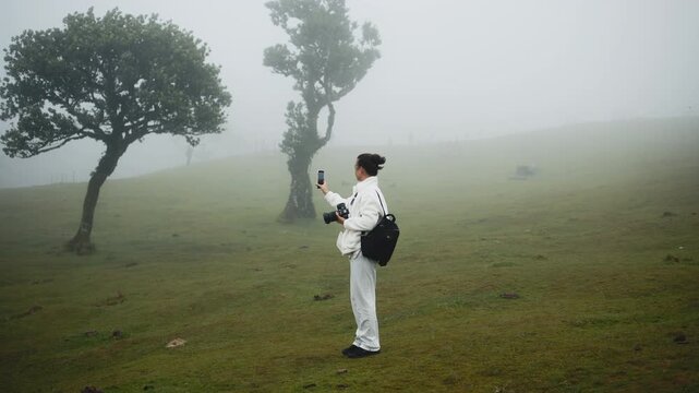 A young woman takes a photo with her phone in foggy nature. Concept capturing moments in misty landscapes.