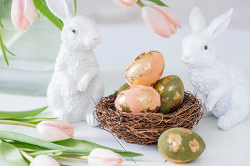 Beautiful Easter greeting card. A bouquet of delicate tulips, Easter bunnies, and golden-patterned eggs on a table in a white kitchen interior.