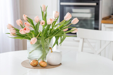 Beautiful Easter greeting card. A bouquet of delicate tulips, a chicken, and eggs with a golden pattern on a table in a white kitchen interior.