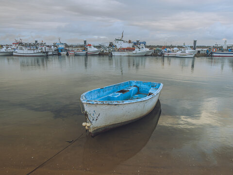 View of a lone blue and white boat resting on the wet sand, reflecting in the water with larger boats docked in the distance, Punta Umbr&iacute;a, Andalusia, Spain.